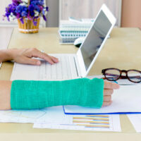 working woman with green cast on arm working on laptop in office, focus on broken hand