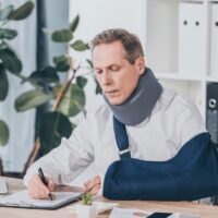 worker in neck brace and arm bandage sitting at table and carefully writing on document with pen in office, compensation concept