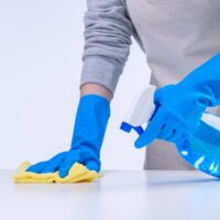 Young woman housekeeper is doing cleaning white table in apron with blue gloves, spray cleaner, wet yellow rag, close up, copy space, blank design concept. Young woman housekeeper is doing cleaning white table in apron with blue gloves, spray cleaner, wet yellow rag, close up, copy space, blank design concept.