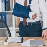cropped view of worker with broken arm in bandage standing near table in office, compensation concept