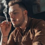Man blowing out puffs of smoke while smoking weed hand rolled cigarette at home