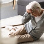 senior woman with migraine sitting on carpet and touching forehead with hand