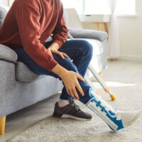 Close-up of handicapped man in casual clothes with leg in cast with crutch sitting on sofa at home in rehabilitation and recovering after having fractured limb. Recovery, accident and rehab concept.