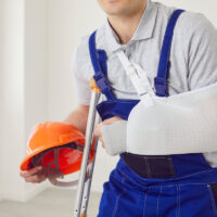 Croped shot of repairman with arm bandage and crutches standing after accident at workplace and holding hard hat. Male builder or foreman in workwear uniform got injured during construction. Croped shot of repairman with arm bandage and crutches standing after accident at workplace and holding hard hat. Male builder or foreman in workwear uniform got injured during construction.