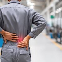 A factory worker holds his sore back, highlighted in red, indicating pain, from injury or strain.
