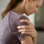 A person in a purple shirt holds their shoulder with a pained expression. The scene suggests discomfort or pain, conveying a sense of vulnerability.