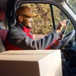 a-delivery-driver-in-a-red-jacket-and-cap-smiles-while-using-a-digital-tablet-inside-a-van